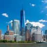 One World Observatory City Cards & Passes (New York) Several modern skyscrapers, including a tall, pointed tower, stand along the waterfront under a clear blue sky.