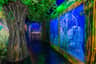 An indoor aquarium with large fish tanks, tree-like structures, lush green foliage, and vibrant lighting in a jungle-themed setting.